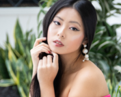 A woman sits outdoors in a pink off-shoulder dress with large pearl earrings