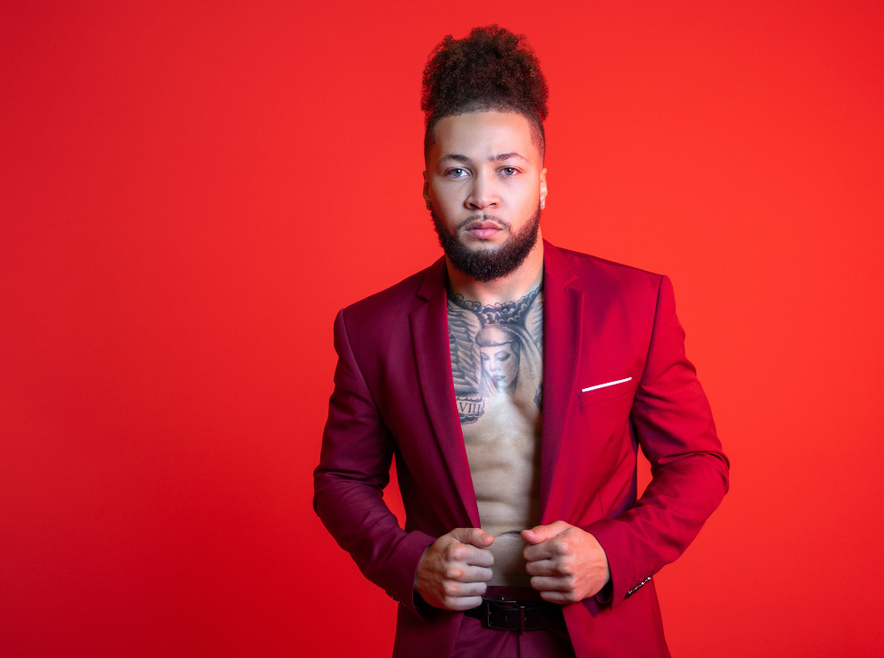 A light-eyed, dark-haired bearded man with a tattooed chest poses in a burgundy suit against a red background