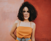 A woman in an orange top stands against a half pink and half red wall