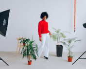 A woman in a red top and white pants stands surrounded by potted plants and studio lights against a white background in a studio