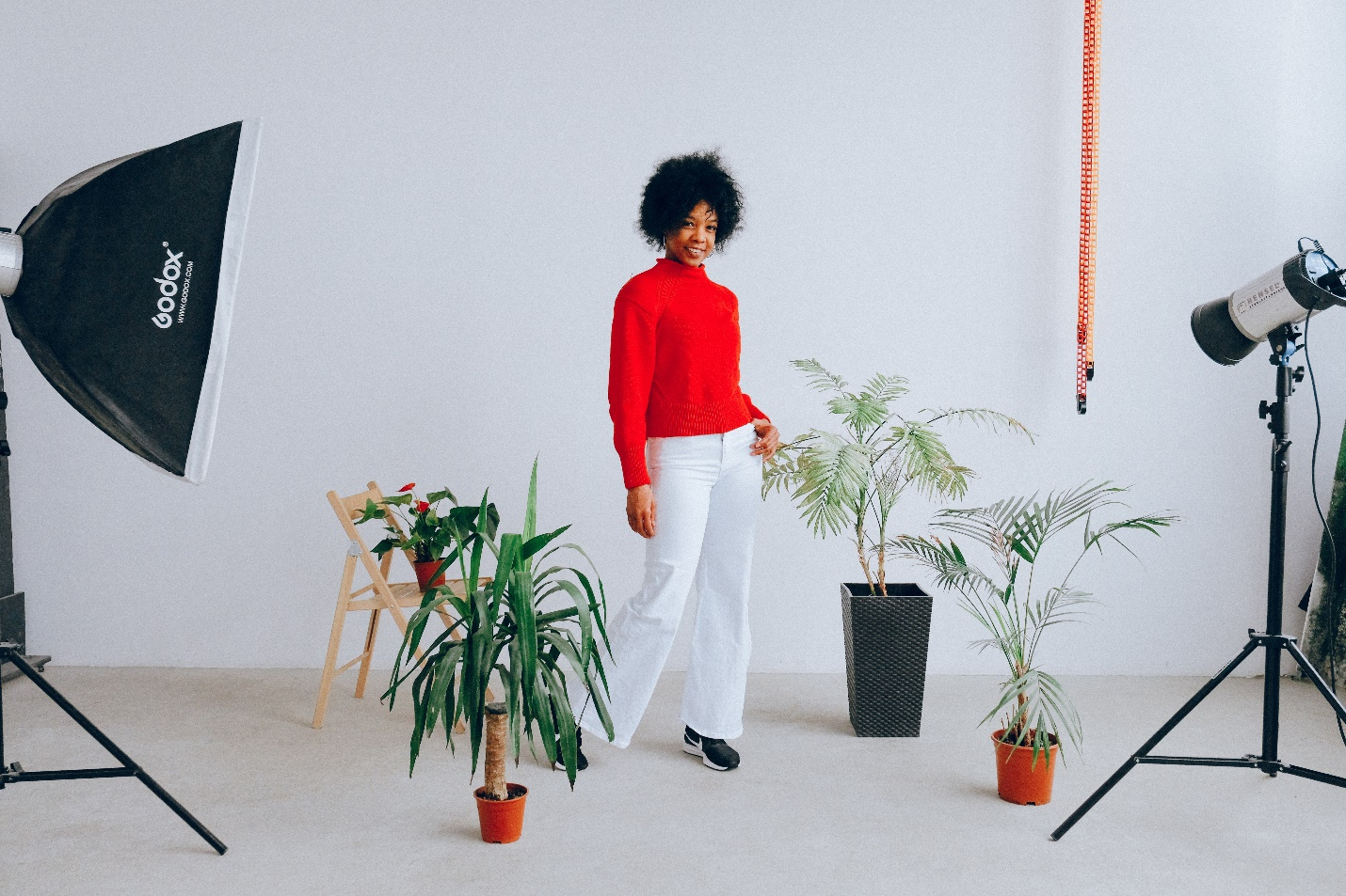 A woman in a red top and white pants stands surrounded by potted plants and studio lights against a white background in a studio
