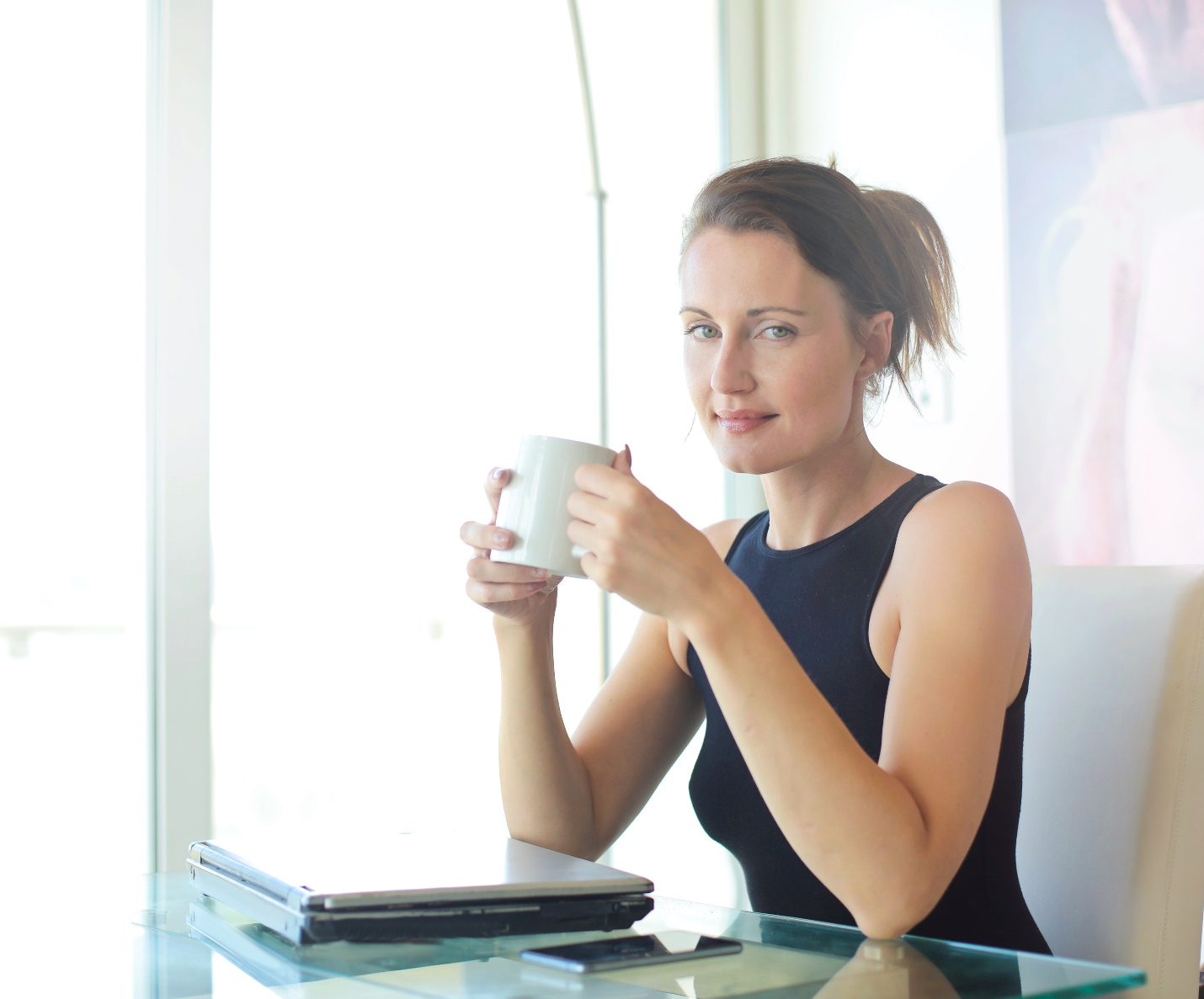 A professional woman sits at her desk with a laptop and phone holding a white mug in her hands